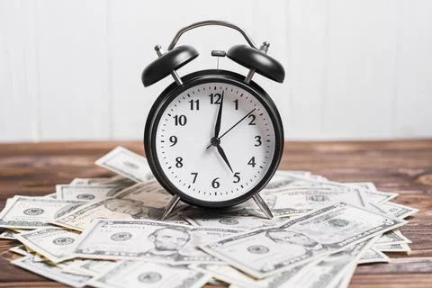 Alarm clock on spread currency notes over wooden textured background. High Stock Photos