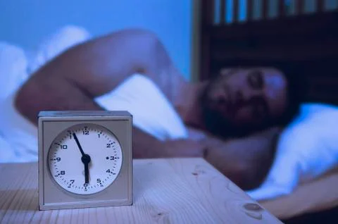 Alarm clock on table, man sleeping in background Stock Photos