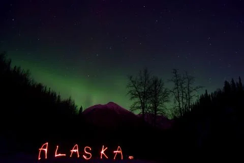 Alaska written in red light in front of a mountain landscape with aurora Stock Photos