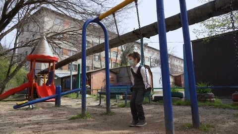 Alone kid in a school suit in face mask sits on swing on an empty playground Stock Footage 127831531