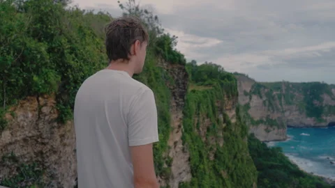 Alone stooped man in a white T-shirt stands on the edge of a cliff and looks at Video stock 249867464