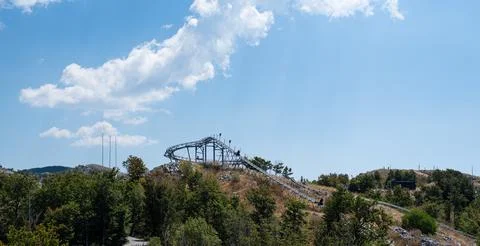 Alpine coaster track winds down a grassy, forested hillside under a blue sky Stock Photos