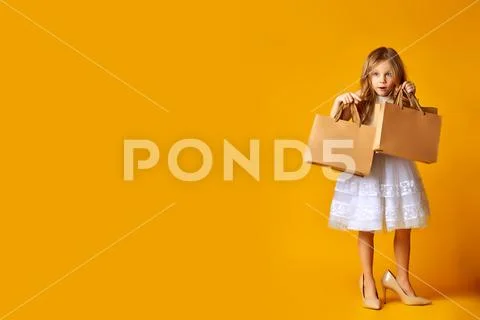 Amazed attractive child in dress and big shoes holding shopping bags on yello Stock Photos