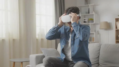 Amazed hipster man using his VR headset while sitting on sofa at home in the Stock Footage 169320662