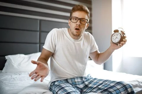 Amazed scared young man on bed this morning. He look on camera and hold clock 写真素材