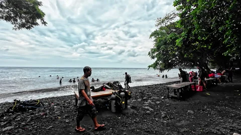 Amed Bali beach with divers, black shore, ocean waves, and cloudy tropical sky Stock Footage 336033891