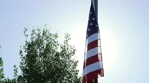 American flag hangs listless as bright sun flares up to reveal and obscure it Stock Footage 202223507