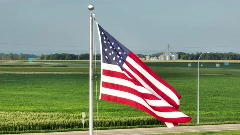 American flag waving in front of corn field and grain elevators in midwest Stock Footage 247675525