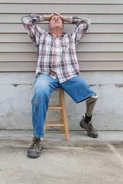 Amputee man sitting on a stool, prosthetic leg out, hands on head looking up Stock Photos