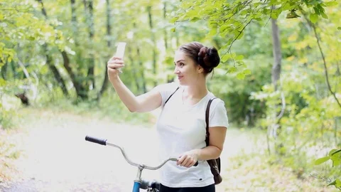 An amusing girl with a backpack and a bicycle makes selfie photo on her phone. Stock Footage 79479118