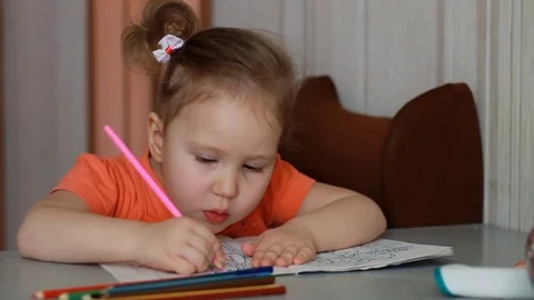 An amusing pretty girl sits at a table and draws in multicolored pencils. Child Stock Footage 82033055