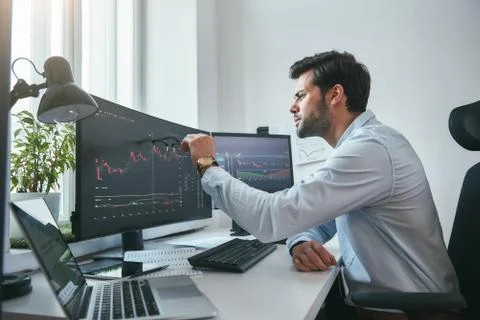 Analyzing data. Thoughtful young trader is pointing at graphs on computer screen Foto stock