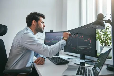 Analyzing data. Thoughtful young trader is pointing at graphs on computer screen Foto stock