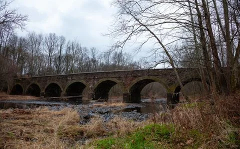 An ancient moss-covered stone arch bridge crosses a quiet river surrounded .. 스톡 사진