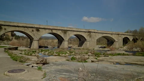 Ancient stone arch bridge crossing a low water river with rocks and dry veg.. Stock Footage 335462376