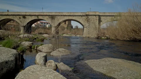 Ancient stone arch bridge spanning a river with rocks and natural vegetation Stock Footage 335463136