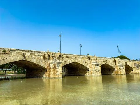 An ancient stone bridge with multiple arches spans over tranquil water, surro Stock Photos