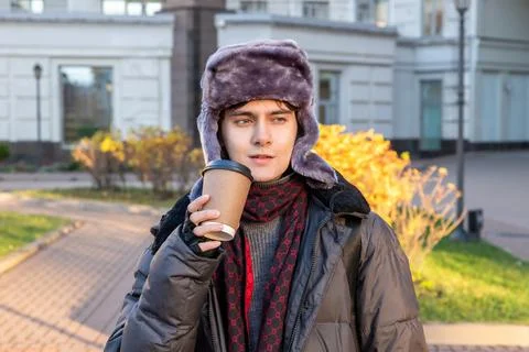 Androgynous man in fur hat drinking takeaway coffee outdoors. Young stylish.. Stock Photos