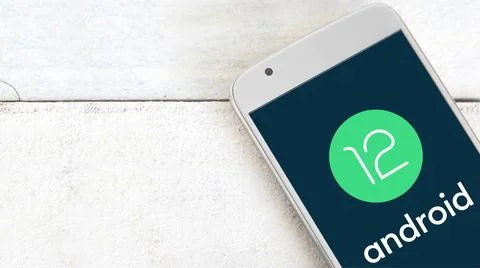 Android 12 logo on a screen of a mobile on a white wooden table. Stock Photos