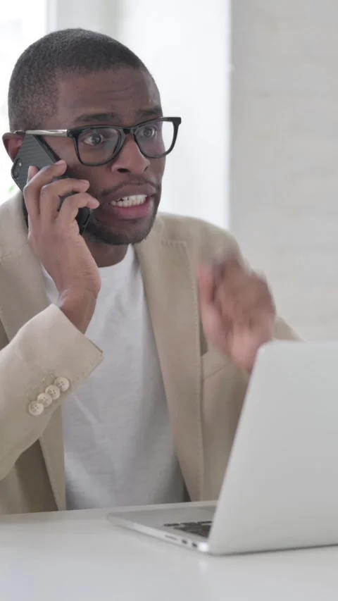 Angry African Man Talking on Smartphone while using Laptop in Office, Vertical Vídeos de archivo 337350098