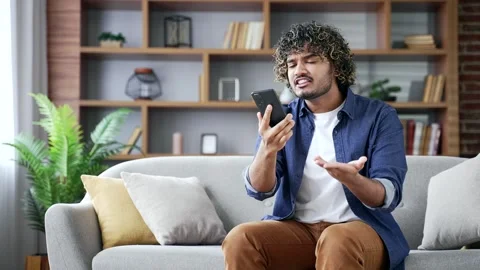 Angry excited man talking arguing on smartphone sitting on sofa in apartment  Stock Footage 296757892