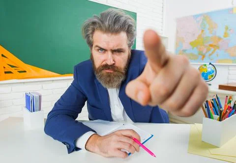 Angry male teacher in classroom pointing finger. Serious bearded man in suit  Stock Photos