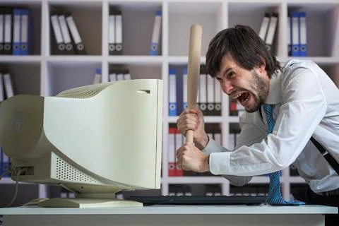 Angry man is destroying old computer with baseball bat in office. Stock Photos