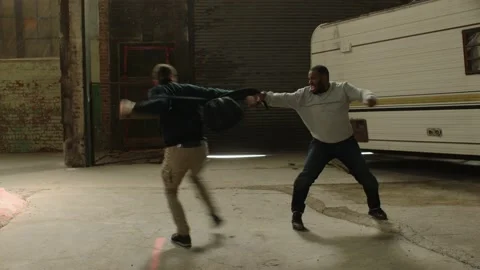 Angry man drop kicks weaker man while fighting over backpack in front of an aban Video stock 199457269