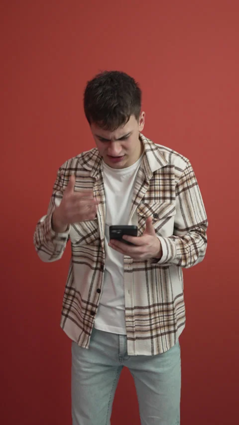Angry man use mobile phone stand in front red background studio shot Video stock 300789644