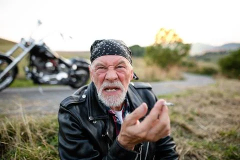 Angry senior man traveller with motorbike in countryside, smoking. Stock Photos