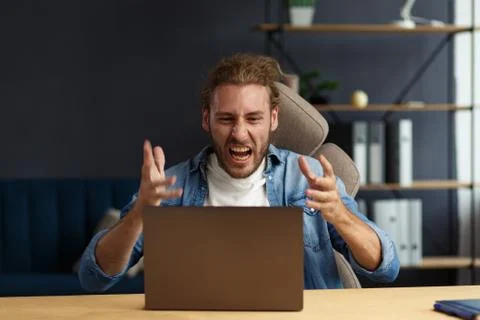Angry young handsome curly man using laptop. Stressed freelancer lost data on Stock Photos