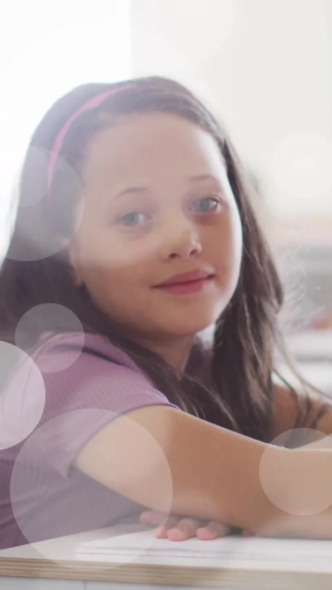Animation of bokeh light spots over happy schoolgirl sitting at desk in class Stock Footage 323898469