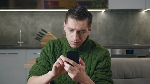 Annoyed man uses a smartphone while sitting on the couch at home. The young male Stock Footage 197357899