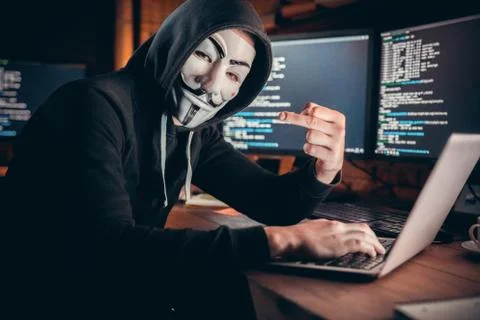 Anonymous hacker looking at camera showing fuck hacking on laptop Stock Photos