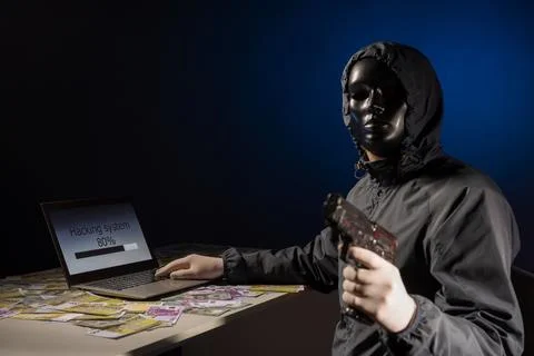 Anonymous hacker programmer uses a laptop holding a gun in his hand to hack.. Stock Photos