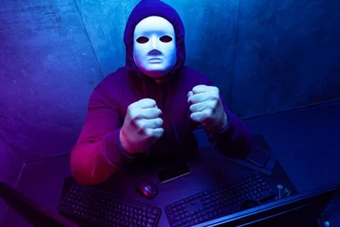 Anonymous hacker wearing face mask working on computer in dark room Stock Photos