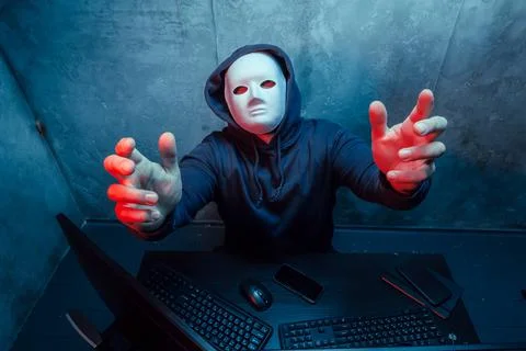 Anonymous hacker wearing face mask working on computer in dark room Stock Photos