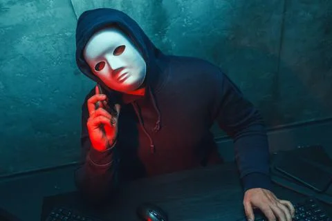 Anonymous hacker wearing face mask working on computer in dark room Stock Photos