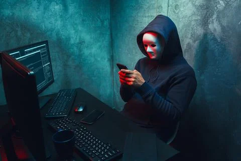 Anonymous hacker wearing face mask working on computer in dark room Stock Photos