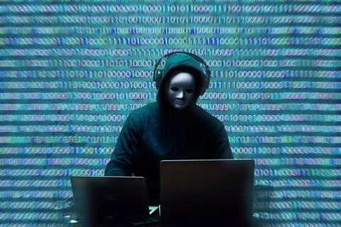 Anonymous hacker wearing face mask working on computer in dark room Stock Photos