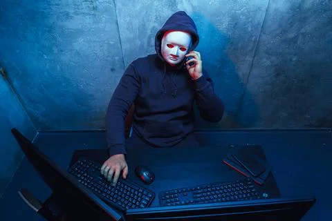 Anonymous hacker wearing face mask working on computer in dark room Stock Photos