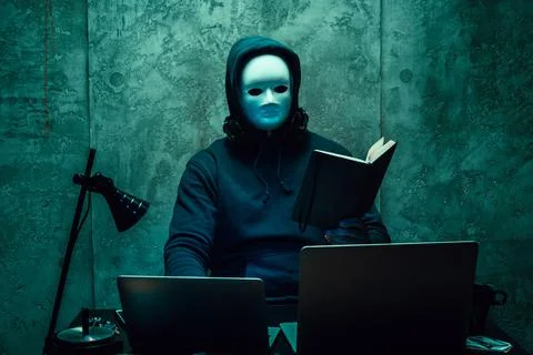 Anonymous hacker wearing face mask working on computer in dark room Stock Photos