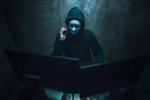 Anonymous hacker wearing face mask working on computer in dark room Stock Photos