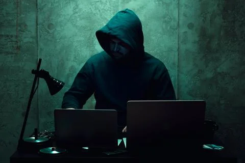 Anonymous hacker wearing face mask working on computer in dark room Stock Photos
