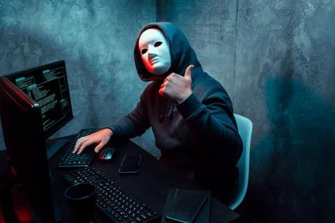 Anonymous hacker wearing face mask working on computer in dark room Stock Photos