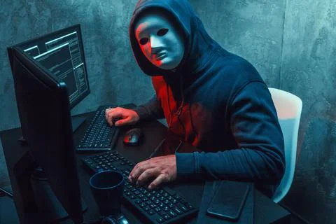 Anonymous hacker wearing face mask working on computer in dark room Stock Photos