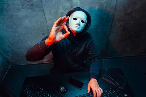 Anonymous hacker wearing face mask working on computer in dark room Stock Photos