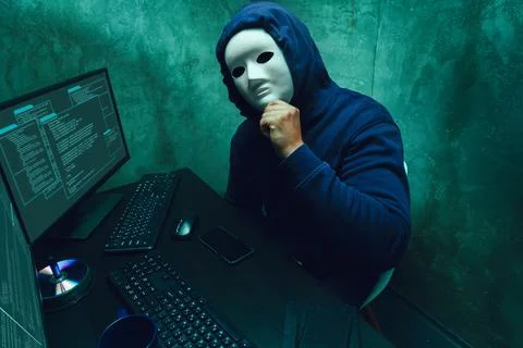 Anonymous hacker wearing face mask working on computer in dark room Stock Photos