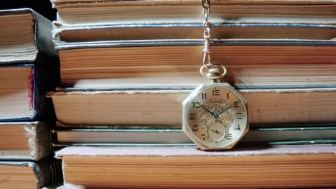 Antique gold pocket watch on chain hanging against background of stacks of books Stock Footage