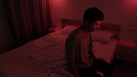 Anxious man sitting alone on the edge of his bed, lost in worried thoughts and Vidéo 347298024